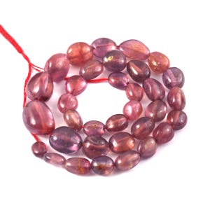 Peut inclure: Un collier de perles en pierre précieuse polies de forme ovale, aux teintes roses et violettes, disposées en cercle. Les pierres lisses et translucides varient en taille et en intensité, allant du magenta clair au violet profond, sur un fil rouge.