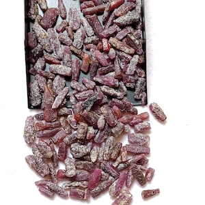 Op de afbeelding: Een verzameling ruwe, ongepolijste robijnkristallen in verschillende tinten dieprood en roze. De stenen hebben een ruw, getextureerd oppervlak met enkele grijze en witte minerale insluitingen en liggen in en morsen uit een zwarte tray.