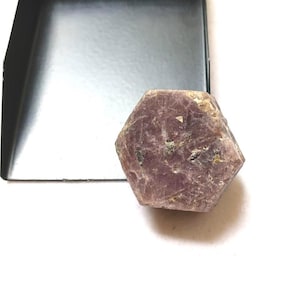 Peut inclure: Un spécimen minéral hexagonal brut de couleur violet poussiéreux avec une surface cristalline texturée. La pierre présente des inclusions naturelles et des variations de couleur, allant du mauve clair au prune profond, placée près d'un objet en métal noir.