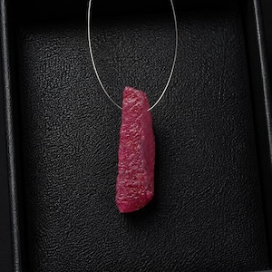 Peut inclure: Un pendentif en pierre précieuse brute d'un rouge rosé profond, suspendu à un fil fin de couleur argentée. La pierre présente une surface rugueuse, texturée et cristalline avec une forme allongée et irrégulière, révélant des facettes naturelles.