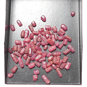 May include: A collection of small, raw, pinkish-red gemstone crystals with irregular, faceted surfaces scattered across a black tray. The stones vary in shape and size, displaying a translucent quality and a natural, unpolished texture throughout.