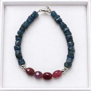 Puede incluir: Una pulsera compuesta por una hilera de pepitas de piedras preciosas en bruto de color azul oscuro y cuatro cuentas de piedras preciosas rojas lisas de forma ovalada. La pulsera incluye pequeñas cuentas metálicas plateadas y un cierre de mosquetón.