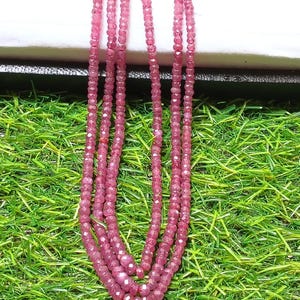 Può includere: Una collana a più fili composta da piccole perle di pietre preziose sfaccettate di colore rosa intenso. I fili sono disposti in un anello a cascata, mettendo in risalto la consistenza uniforme e riflettente delle singole pietre del gioiello.