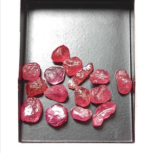 Puede incluir: Una colección de catorce piedras preciosas rojas en bruto y sin pulir esparcidas sobre una bandeja rectangular de metal negro. Las piedras presentan formas irregulares variadas, texturas de translúcidas a opacas y tonos carmesí profundos.