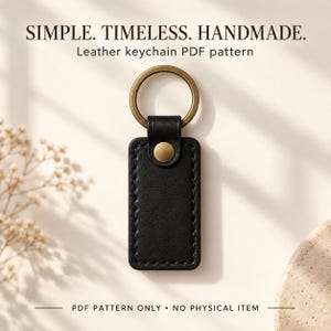 Hand holding a black rectangular leather keychain by its brass ring, showing stitched edges and a brass snap, with “Made by You” text.