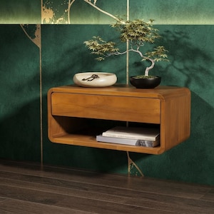May include: A wall-mounted wooden floating nightstand with rounded corners, featuring a single top drawer and an open lower shelf. On the surface sits a cream-colored ceramic bowl containing keys and a small potted bonsai tree in a black pot.