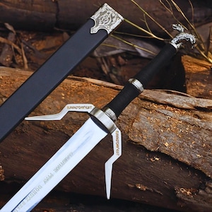 May include: A silver-toned steel sword with a black wrapped handle, a wolf-shaped pommel, and a crossguard featuring angular prongs. The blade is engraved with text, and the sword is accompanied by a black scabbard with ornate silver metal fittings.
