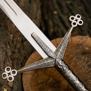 Puede incluir: Una espada de metal plateado con una hoja larga y recta que presenta un canal central. La empuñadura muestra grabados intrincados de volutas, un relieve de león y una guarda que termina en formas de trébol de cuatro círculos.