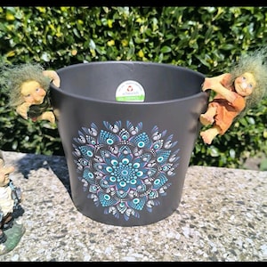 Könnte beinhalten: Ein matter, dunkelgrauer Pflanztopf mit einem zentralen Mandala-Design aus kleinen türkisfarbenen, weißen und violetten Punkten. Zwei kleine Trollfiguren mit grünem Haar sind am Rand befestigt, eine auf jeder Seite, und ein Aufkleber ist innen.