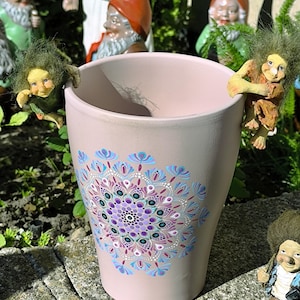 Könnte beinhalten: Ein hellrosa konischer Pflanztopf mit einem handgemalten Mandala-Muster in Lila-, Blau- und Weißtönen. Zwei kleine Trollfiguren mit wildem grünem Haar sind am Rand befestigt und greifen jeweils von einer Seite über den Topfrand.