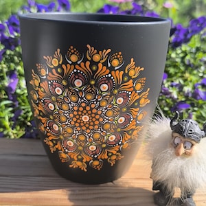 Könnte beinhalten: Ein mattschwarzer Becher mit einem handgemalten Mandala-Design in Orange-, Gelb- und Weißtönen. Das komplexe Muster besteht aus zahlreichen kleinen Punkten, die in konzentrischen Kreisen angeordnet sind. Eine kleine Wikingerfigur steht daneben.