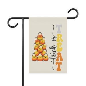 May include: A rectangular garden flag featuring a stack of nine cartoon candy corn pieces with smiling faces. The text "trick or TREAT" is printed vertically in a wavy, multi-colored font. The flag hangs from a black metal garden stake.