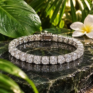 Puede incluir: Una pulsera de tenis en tono plateado con una hilera continua de piedras redondas, transparentes y facetadas engastadas en un marco metálico. La pulsera incluye un cierre de caja seguro y se muestra en forma circular sobre una superficie oscura.