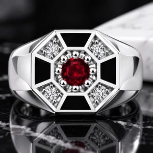 Op de afbeelding: Een zilverkleurige ring met een centrale rondgeslepen rode edelsteen omringd door kleine zilveren bolletjes. De ringkop heeft een achthoekig geometrisch ontwerp met vier heldere rondgeslepen stenen en vier zwarte, met email gevulde driehoeken.