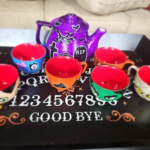 May include: A purple teapot with a spooky design and six teacups with different spooky designs. The teacups and teapot are on a black tray with a white ouija board design. The text on the board reads "1234567890 GOOD BYE".