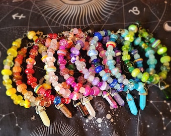 May include: A collection of colorful beaded necklaces in a rainbow of hues, including yellow, red, pink, purple, blue, and green. Each necklace features a unique combination of beads and a crystal pendant. The necklaces are displayed on a black surface.