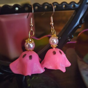 May include: A pair of pink ghost earrings with gold-colored hooks. Each earring features a pink ghost with two dark brown eye holes and a pearl bead with gold-colored accents. The earrings are displayed against a dark background.