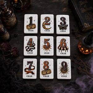 May include: Nine rectangular white tiles arranged in a grid, each featuring a large black number from one to nine and the word CRAK. The designs include an admit one ticket, crescent moon, moth, snake, sunburst, circus tent, ribbon, lucky 8 ticket, and starburst.