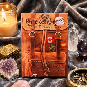 May include: A rectangular tarot training deck box featuring a brown leather backpack illustration with two straps, buckles, and travel stickers including a Canadian flag. The text reads Broke Biker Tarot Training Deck by Kat Dehring and Taryn Masterson.