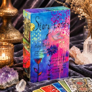 Può includere: Una scatola rettangolare di Story Cards dell'artista Cathy Nichols con un vivace design astratto multicolore, fiori dipinti, un uccello e testo. La scatola include 70 carte dipinte a mano e un libretto di istruzioni.