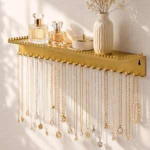 Könnte beinhalten: Ein goldfarbenes Wandregal zur Schmuckaufbewahrung mit einer Ablagefläche für Accessoires und einer Reihe von Haken darunter. Mehrere goldene Ketten und Halsketten mit verschiedenen Anhängern und Perlen hängen an den Haken, neben zwei Parfümflakons.