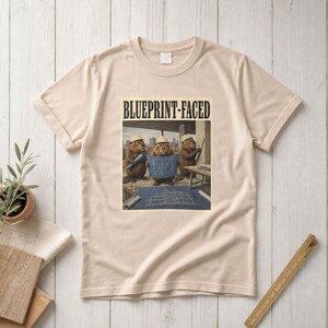Könnte beinhalten: Ein beiges Kurzarm-T-Shirt mit einem Grafikdruck von drei Bibern, die Bauhelme tragen. Die Biber halten Baupläne auf einer Baustelle. Der Text "BLUEPRINT-FACED" ist in schwarzer Schrift über dem Bild aufgedruckt.