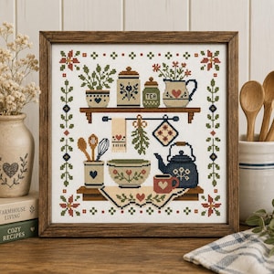 Peut inclure: Une broderie au point de croix encadrée représentant une scène de cuisine avec deux étagères en bois contenant un pot à thé, une carafe à fleurs, un bol, une théière bleu foncé et une tasse. Le motif inclut des torchons et ustensiles.