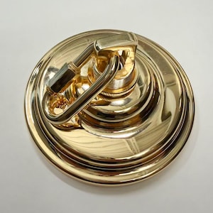 May include: A polished gold-tone metal ceiling canopy kit featuring a circular mounting plate with a stepped edge design. A matching gold-tone carabiner hook is securely attached to the center threaded loop, providing a functional hardware component.