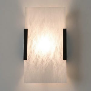May include: A rectangular wall sconce featuring a translucent white panel with a marbled texture. The light is illuminated from within, casting a soft glow. Two vertical matte black metal brackets secure the panel on the left and right sides.