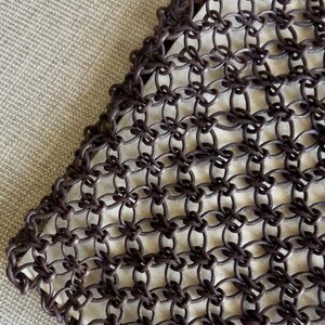May include: A dark brown garment featuring an intricate, open-work chainmail-style construction. The design consists of numerous interconnected circular rings linked together to form a repeating grid pattern, layered over a solid light-colored fabric.