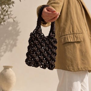 May include: A dark brown handbag featuring a complex, open-weave macrame construction made from thick, knotted cord. The bag has two integrated circular handles formed by the same knotted material, creating a cohesive, textured, and structured design.