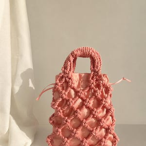 May include: A peach-colored handbag featuring a knotted rope overlay in a lattice pattern. The bag has two thick, braided rope handles and a drawstring closure made of matching satin fabric, creating a textured and layered appearance.
