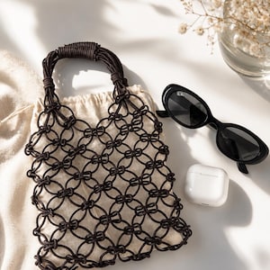 May include: A dark brown macrame handbag featuring a circular knotted pattern and a matching handle, paired with black oval sunglasses and a white wireless earbud charging case. The bag has a light beige fabric lining visible through the knots.