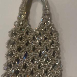 May include: A silver-toned handbag featuring an intricate, open-weave lattice pattern constructed from small, reflective, metallic beads. The bag has a structured, rounded top handle and a rectangular body with a shimmering, textured surface.