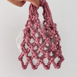 Può includere: Una borsa a mano in macramè color rosa polvere con un design a corda annodata a motivo romboidale. La borsa include una pochette interna bianca traslucida con coulisse visibile attraverso la struttura a corda aperta e due manici integrati.