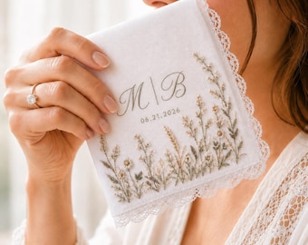 May include: A white fabric handkerchief featuring delicate embroidery of wildflowers in muted green and brown tones. The centre displays the embroidered initials M | B above the date 21.06.2026. The edges are finished with a white scalloped lace trim.
