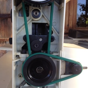 Puede incluir: Primer plano de una máquina de coser con una correa verde. La correa envuelve dos poleas, una grande y una pequeña. La máquina es blanca con detalles negros.