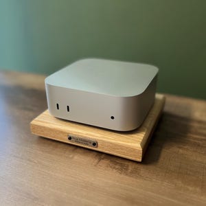 May include: A silver, square-shaped electronic device with rounded corners and two vertical slots and one circular port on the front, resting on a light-colored wooden base featuring a small metal plaque that reads Nua Materia Made in Italy.