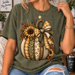 May include: An olive green short-sleeve t-shirt featuring a large graphic of a pumpkin decorated with sunflowers, wheat, and a plaid bow. The pumpkin design includes various patterns like stripes and floral prints in shades of orange, yellow, and green.