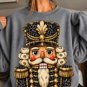 May include: A grey crewneck sweatshirt featuring a large graphic print of a nutcracker. The nutcracker wears a black and grey leopard print uniform with gold rope trim, a matching hat, and has a white beard, mustache, and hair.
