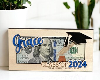 May include: A wooden picture frame with a graduation cap cutout and the text "Class of 2024 Clear Springs High School". The frame has a $100 bill inside and the name "Grace" is written in blue letters.