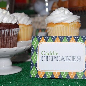 May include: Close-up of cupcakes with white frosting on white pedestals. One cupcake is chocolate, the others are vanilla. A sign in front reads "Caddie CUPCAKES" with a green, blue, and orange diamond pattern.