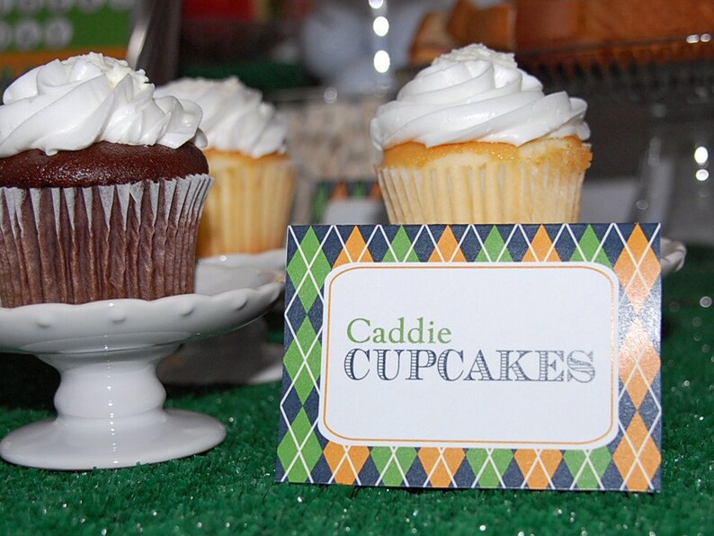 May include: Close-up of cupcakes with white frosting on white pedestals. One cupcake is chocolate, the others are vanilla. A sign in front reads "Caddie CUPCAKES" with a green, blue, and orange diamond pattern.