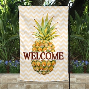 May include: A welcome garden flag with a yellow and white chevron pattern. A watercolor pineapple with green leaves is in the center of the flag. The word "WELCOME" is printed in brown letters below the pineapple.