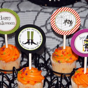 Pode incluir: Cupcakes com cobertura laranja e granulado colorido, decorados com temas de Halloween. As decorações mostram uma aranha, uma bruxa e a frase "Happy Halloween". Os cupcakes são apresentados em um suporte em forma de teia de aranha preta.