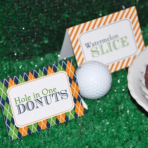 May include: Two table cards with text, "Hole in One DONUTS" and "Watermelon SLICE", sit on a green surface next to a golf ball and a plate with a donut. The cards have a white border and a diamond pattern in green, navy, and orange.