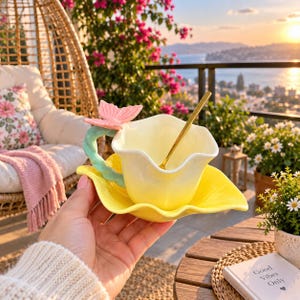 Puede incluir: Un juego de taza y platillo de cerámica amarilla con forma de flor, que presenta un asa en forma de tallo verde claro rematada con una mariposa rosa. Una cuchara de metal dorado descansa dentro de la taza, que tiene un acabado brillante y bordes.
