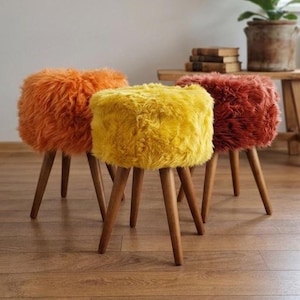 May include: Three round stools with fluffy, long-pile faux fur upholstery in vibrant orange, bright yellow, and deep rust red. Each stool features three tapered, dark brown wooden legs that extend outward from the base to provide stability.