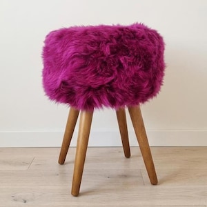 Puede incluir: Un taburete redondo con un cojín de asiento grueso de piel sintética color magenta vibrante. El taburete se apoya sobre cuatro patas de madera de color marrón claro, cónicas y extendidas hacia afuera, que ofrecen una base estable.