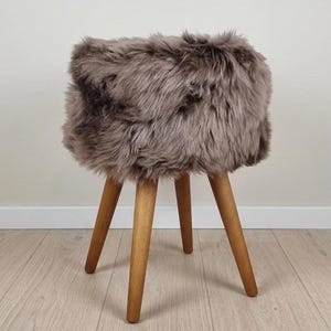 May include: A round stool featuring a thick, plush, taupe-colored faux fur seat cushion supported by four tapered, light brown wooden legs. The stool has a cylindrical top section covered in long, soft-textured fibers and stands on four legs.